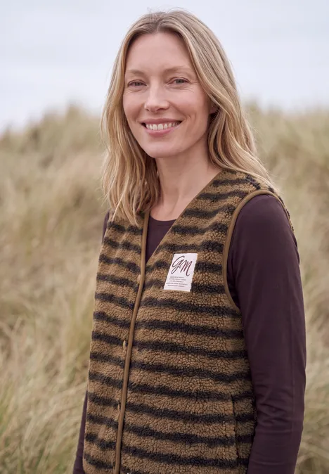 Eine blonde Frau mit gestreifter Weste steht lächelnd vor grasbewachsenen Dünen.