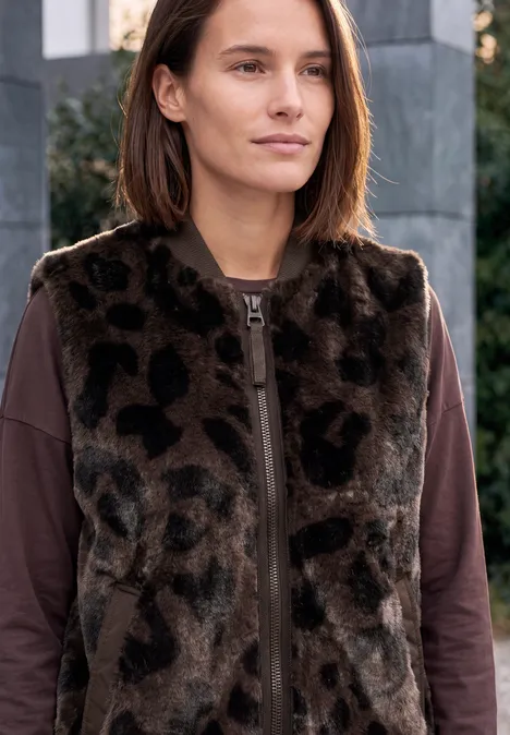 Junge Frau in braunem Langarmshirt und Leopardenfellweste vor moderner Fassade, ruhiger Blick.