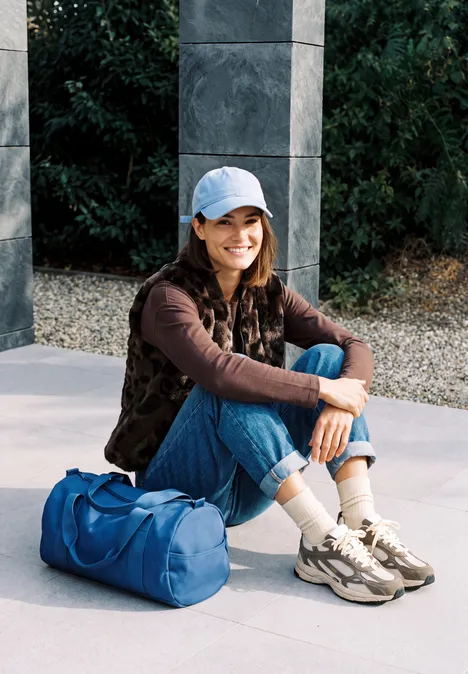 Junge Frau mit Baseballcap und Freizeitkleidung sitzt lächelnd im Freien neben einer blauen Sporttasche.