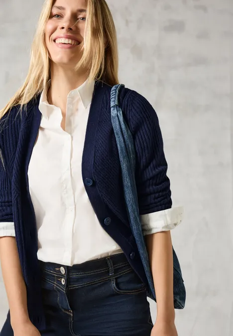 Frau in weißer Bluse und dunkler Strickjacke lächelt, trägt Jeans und eine blaue Tasche.