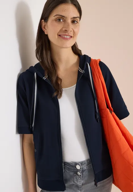 Sweaterjack met korte mouwen en capuchon universal blue Persoon draagt donkerblauwe hoodie over wit topje en oranje tas tegen een lichte achtergrond.