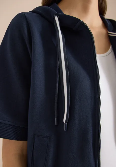 Sweaterjack met korte mouwen en capuchon universal blue Close-up van een borstbeeld met marineblauwe korte-mouwen capuchontrui over een wit topje.