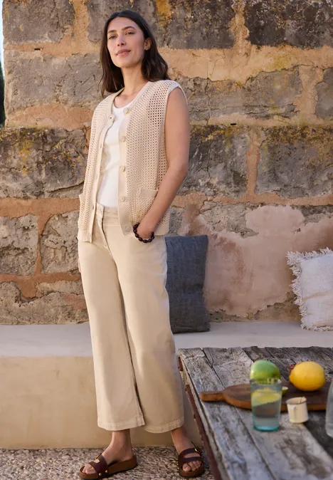 Femme en gilet beige, top blanc et pantalon clair devant un mur en pierre, à côté d’une table rustique avec fruits et verre.