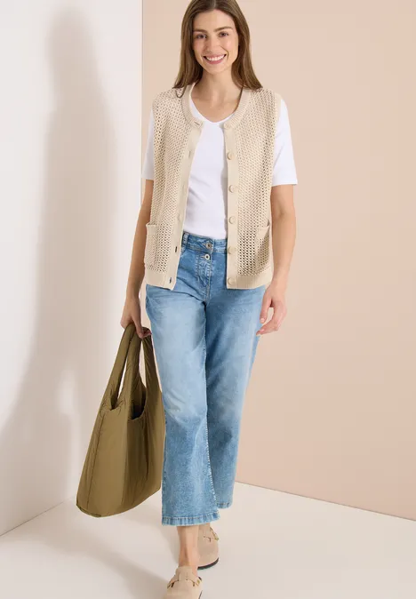 Femme portant un t-shirt blanc, gilet beige, jean bleu, sac olive et chaussures beiges sur fond neutre.