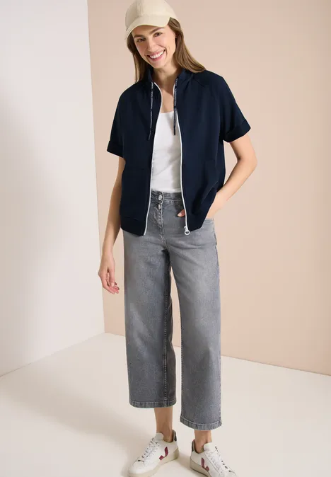 Femme portant un chemisier bleu marine ouvert, un top blanc, un jean gris large 7/8 et des baskets blanches sur fond beige.