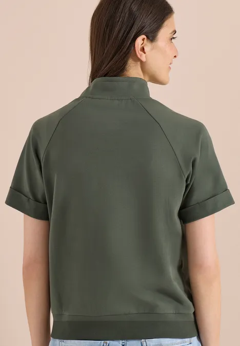 Frau von hinten in olivgrünem Kurzarm-Poloshirt, Haare halboffen, vor neutralem Hintergrund.
