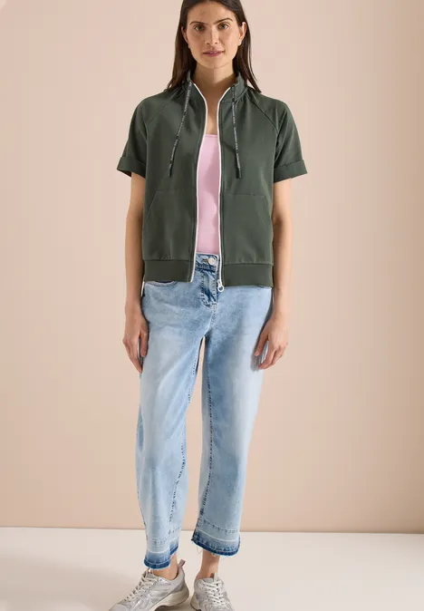 Modell in khakigrüner, kurzärmeliger Kapuzenjacke, rosa Top und hellblauen, umgeschlagenen Jeans.