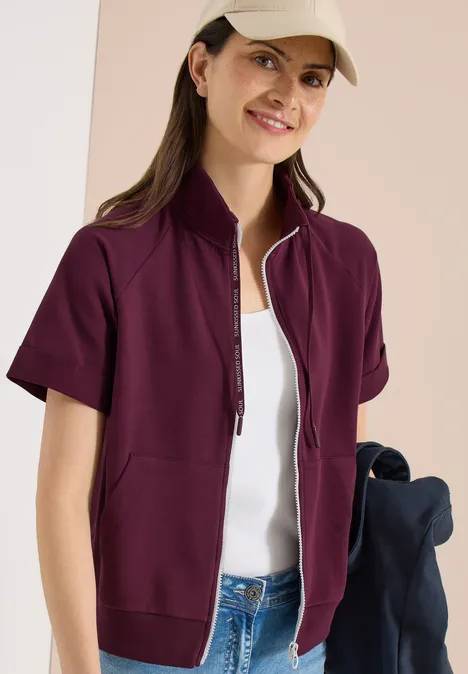 Eine Frau trägt eine kurze, weinrote Jacke mit Reißverschluss über weißem Top und Jeans.