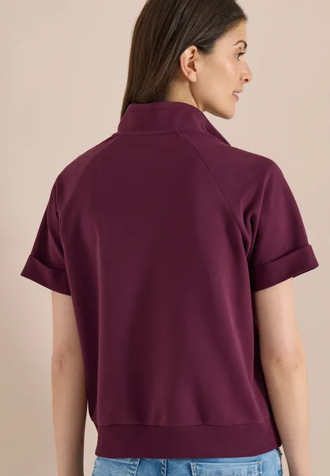 Frau steht mit dem Rücken zur Kamera und trägt ein kurzes, auberginefarbenes Poloshirt mit umgekrempelten Ärmeln.