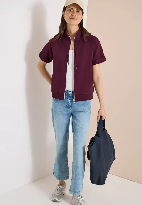 Person mit lila Kurzarm-Jacke über weißem Shirt, blauen Jeans, grauen Sneakern, hält eine dunkelblaue Tasche.