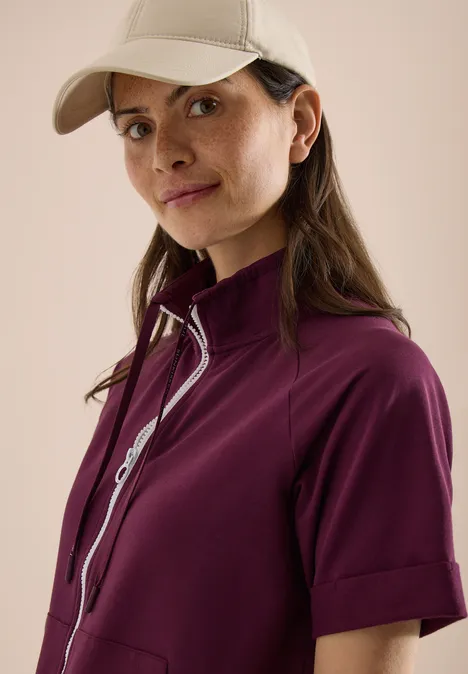 Eine Person in einer lila Jacke und einer beigefarbenen Kappe vor hellem Hintergrund.