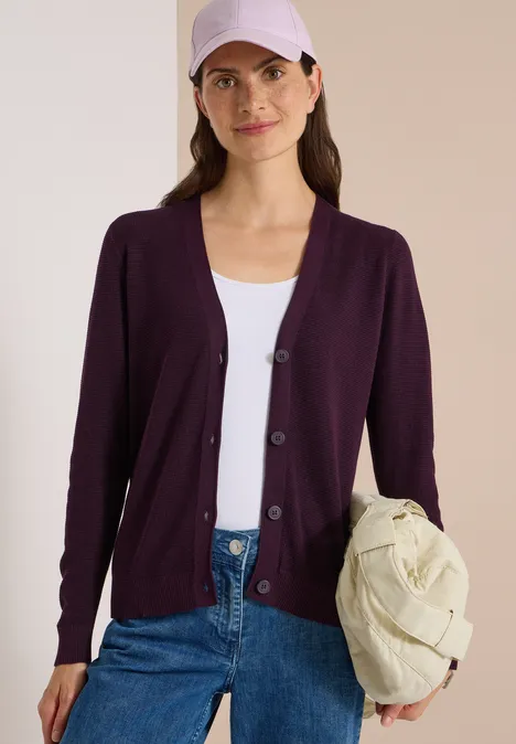 Femme en gilet violet, top blanc et jeans, tenant un sac matelassé clair.