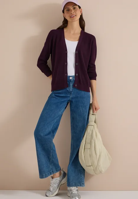 Femme portant un cardigan violet, top blanc, jeans larges, baskets grises, casquette rose et sac matelassé beige.