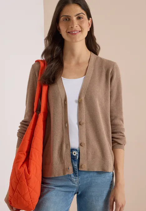 Femme brune souriante, en gilet beige, top blanc, jean bleu et sac matelassé orange.