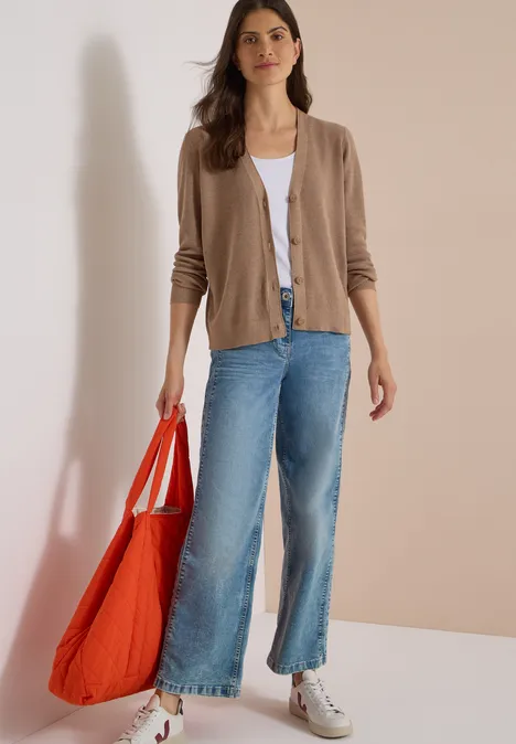 Une femme porte un cardigan beige, un top blanc, un jean flare, des baskets blanches et tient un grand sac rouge.