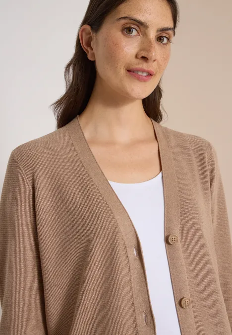 Une femme aux cheveux bruns porte un cardigan beige sur un top blanc et regarde calmement l'objectif.