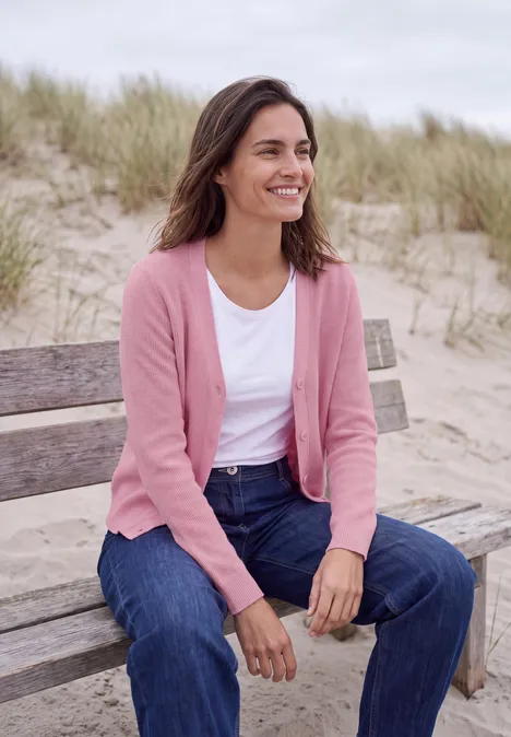 Frau sitzt lächelnd in rosa Strickjacke auf einer Strandbank, Dünen im Hintergrund.