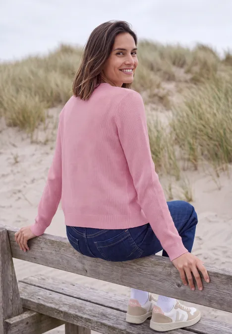 Lächelnde Frau im pinken Pullover und Jeans sitzt auf einer Bank in Dünen und blickt zurück.