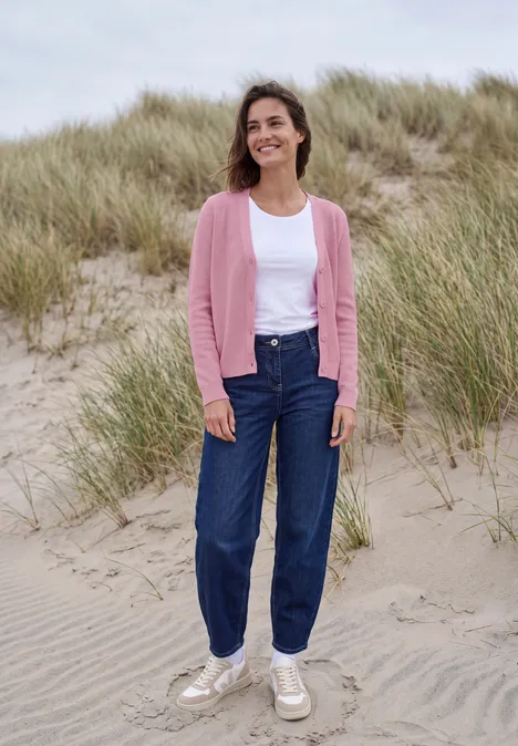Eine lächelnde Frau in Jeans und rosa Strickjacke steht auf sandigen Dünen.