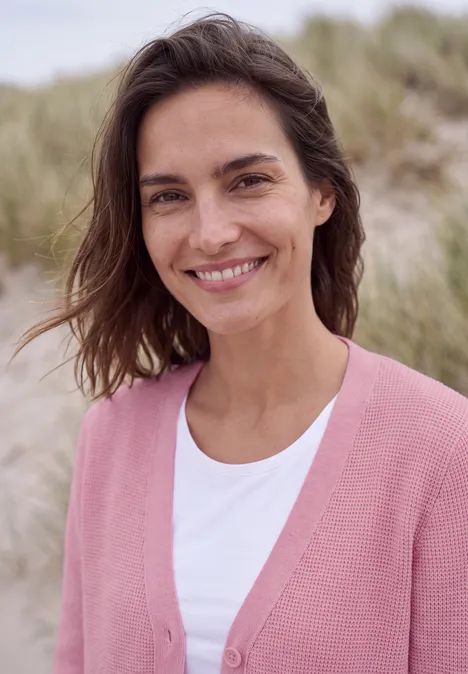 Lächelnde Frau in rosa Strickjacke am Strand mit Dünen im Hintergrund.