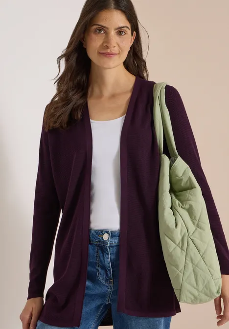 Femme en cardigan violet foncé, top blanc et jean, portant sur l'épaule un grand sac matelassé vert.