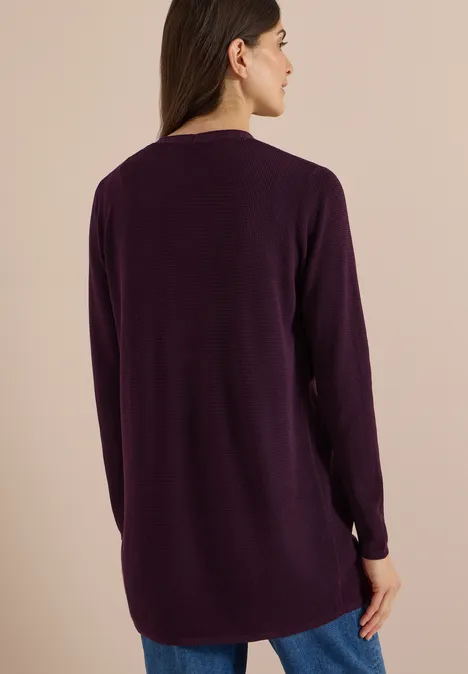 Une femme de dos porte une tunique en maille violette à manches longues devant un fond neutre.