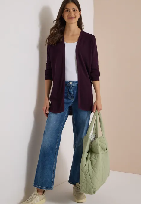 Femme en cardigan violet, top blanc, jean large et baskets, tenant un sac matelassé vert clair.