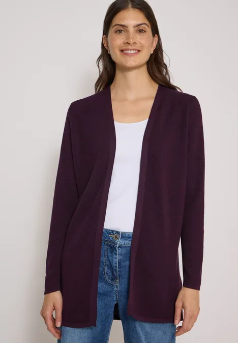Femme en cardigan violet foncé, t-shirt blanc et jean, souriante face à l'appareil photo.