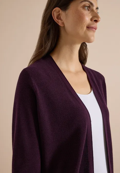 Femme de profil portant un gilet tricoté violet foncé ouvert sur un top blanc, fond neutre.