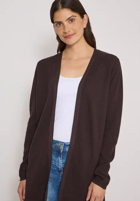 Femme mince portant un gilet marron, un t-shirt blanc et un jean sur fond clair.