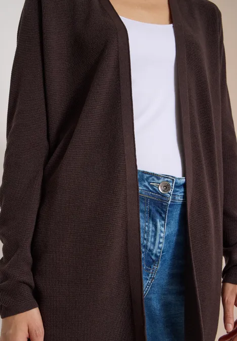 Haut du corps d'une personne portant un cardigan brun ouvert sur un top blanc et un jean bleu.