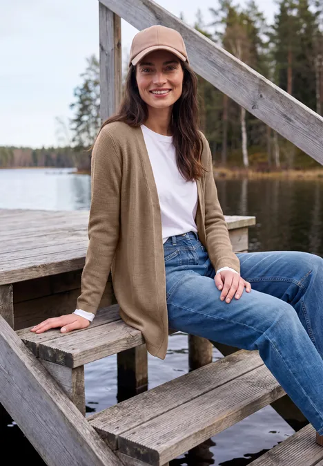 Frau im Beanie, Cardigan und Jeans sitzt auf einem Bootssteg am See, umgeben von Wald und Wasser.