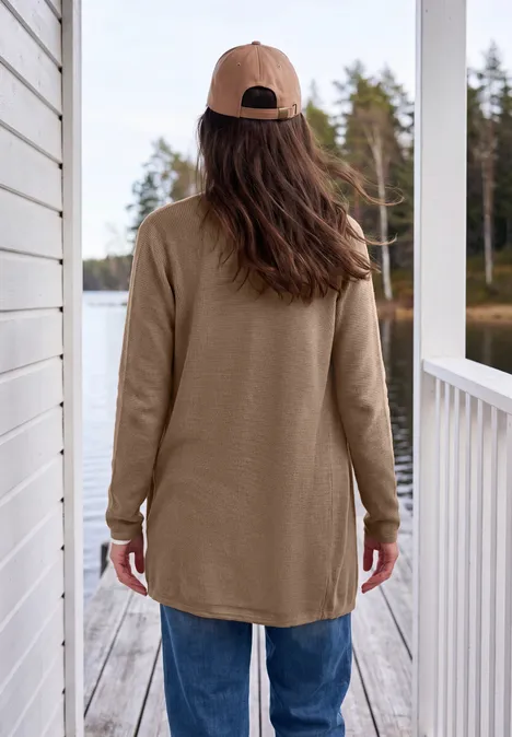 Frau mit brauner Mütze und Longshirt steht auf einer Veranda und blickt auf einen Wald.