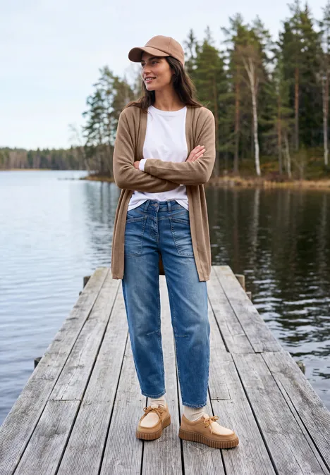 Eine Frau steht auf einem Steg nahe einem Waldsee, trägt Jeans, Cardigan und Mütze.