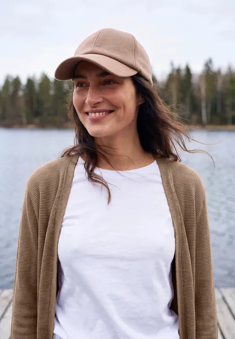 Eine lächelnde Frau in Basecap und weißem Shirt steht entspannt auf einem Steg vor einem ruhigen See, im Hintergrund Wald.