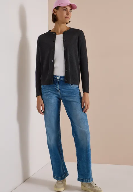 Cardigan boutonné à col rond charcoal grey melange Une femme porte un cardigan noir, un top blanc, un jean bleu et une casquette rose, posant près d’un mur.