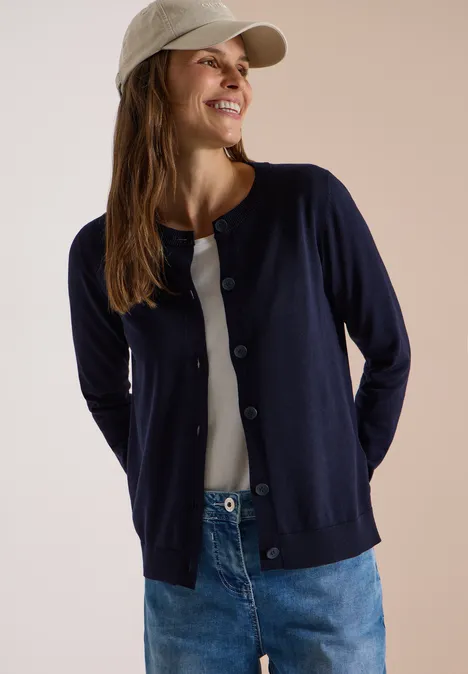Vrouw lacht in spijkerbroek, wit topje, donkerblauw vest en pet tegen een neutrale achtergrond.