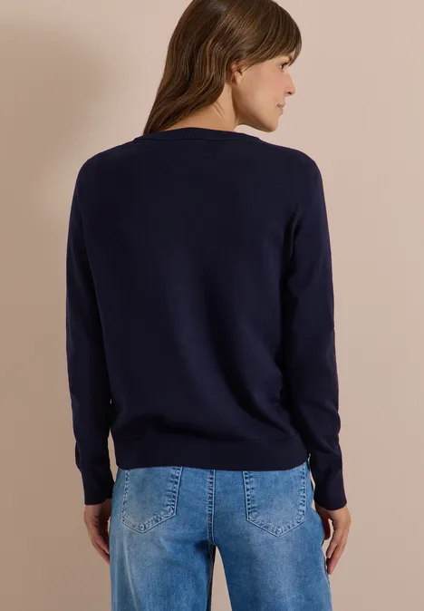 Achteraanzicht van een vrouw met bruin haar in een donkerblauwe trui en lichtblauwe jeans voor een beige achtergrond.