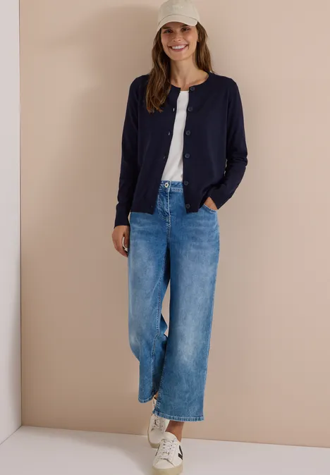 Vrouw in wijde jeans, wit shirt, donkerblauw vest, witte sneakers en pet tegen een beige achtergrond.