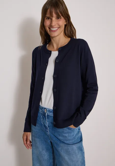 Een glimlachende vrouw draagt een donkerblauwe vest, wit shirt en spijkerbroek.