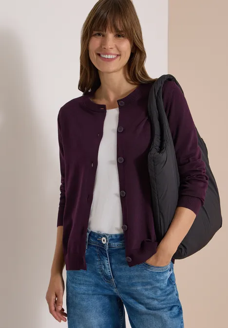 Cardigan à col rond et patte de boutonnage eggplant red Eine lächelnde Frau trägt eine lila Strickjacke, weißes Shirt, blaue Jeans und eine schwarze Schultertasche.