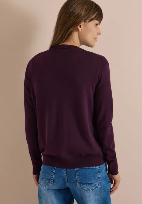 Cardigan à col rond et patte de boutonnage eggplant red Frau von hinten in lila Pullover und Jeans vor beigem Hintergrund.