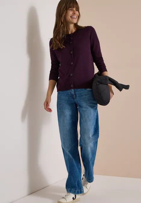 Cardigan à col rond et patte de boutonnage eggplant red Frau in lila Strickjacke und Jeans hält Jacke, geht lächelnd vor hellem Hintergrund.