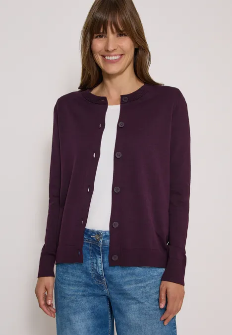 Cardigan à col rond et patte de boutonnage eggplant red Frau in lila Strickjacke, weißem Shirt und blauen Jeans lächelt.