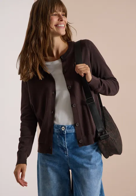 Vrouw in spijkerbroek, wit shirt en bruin vest, met schoudertas, glimlacht voor een neutrale achtergrond.