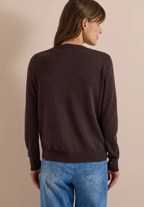 Vrouw van achteren in een bruine trui en blauwe spijkerbroek tegen een neutrale achtergrond.