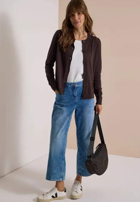 Vrouw met bruin haar in een wit T-shirt, een bruine jas, wijde jeans, witte sneakers en een zwarte tas.