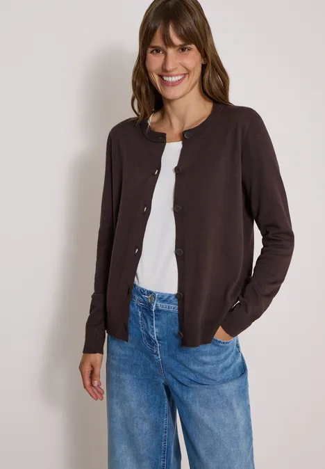 Een lachende vrouw draagt een bruine cardigan, een wit shirt en een blauwe jeans.