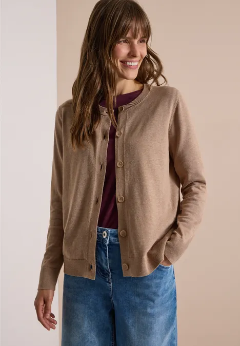 Vrouw lacht en draagt een beige vest, bordeaux top en blauwe jeans, handen in de zakken.