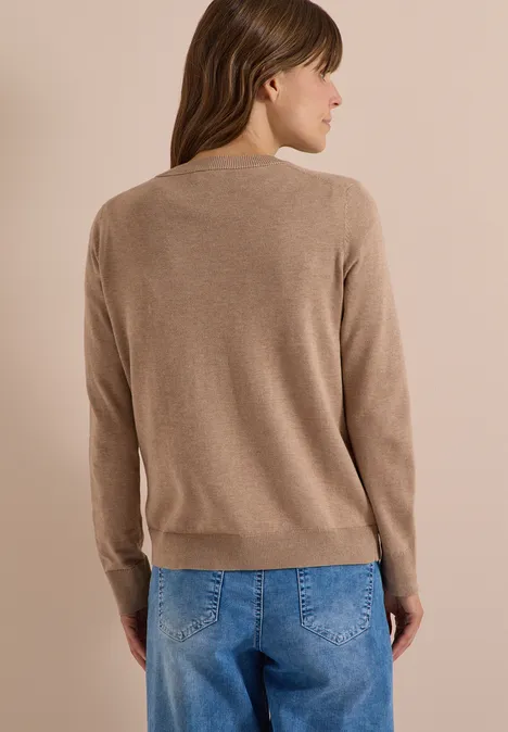 Een vrouw staat met haar rug naar de camera in een beige trui en blauwe jeans.